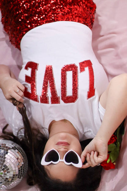 Person wearing a white shirt with red sequin letters spelling 'LOVE' and heart-shaped sunglasses, holding a disco ball.
