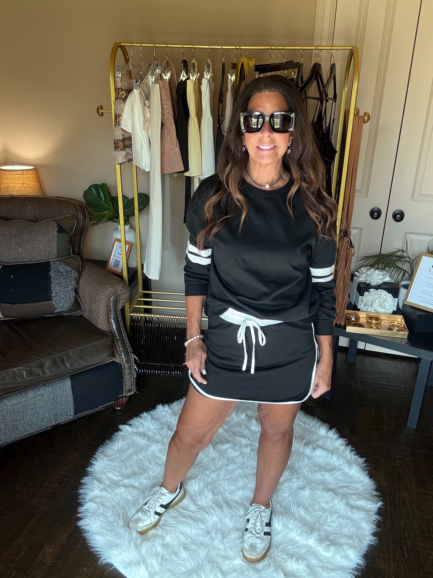 Woman in a black outfit with white drawstrings standing on a white rug in a room with a clothing rack.