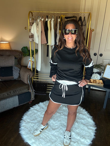 Woman in a black outfit with white accents standing in a room with a clothing rack and sofa.