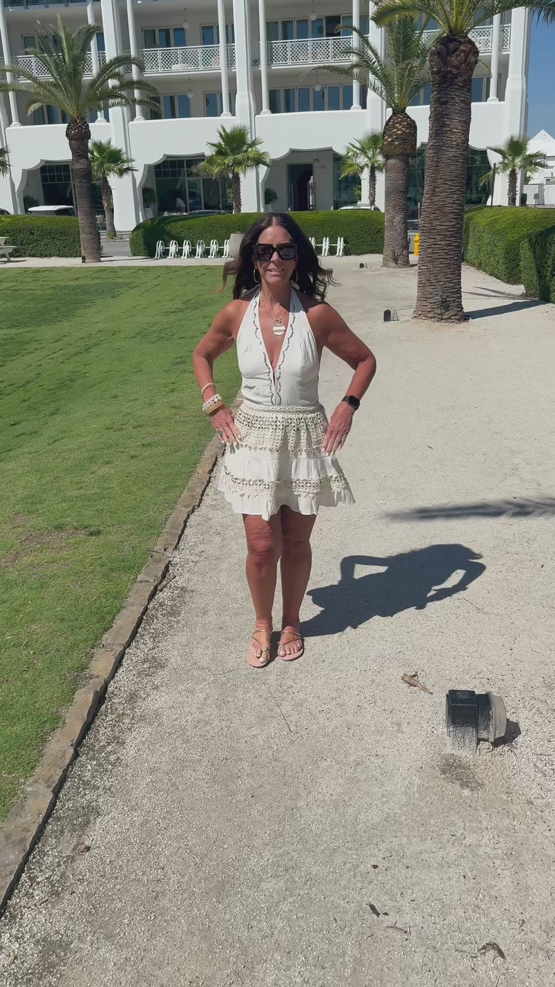 Woman walking on a path with white buildings and palm trees in the background and she is modeling a cream halter dress  with gold accents