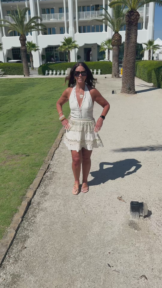 Woman walking on a path with white buildings and palm trees in the background and she is modeling a cream halter dress  with gold accents