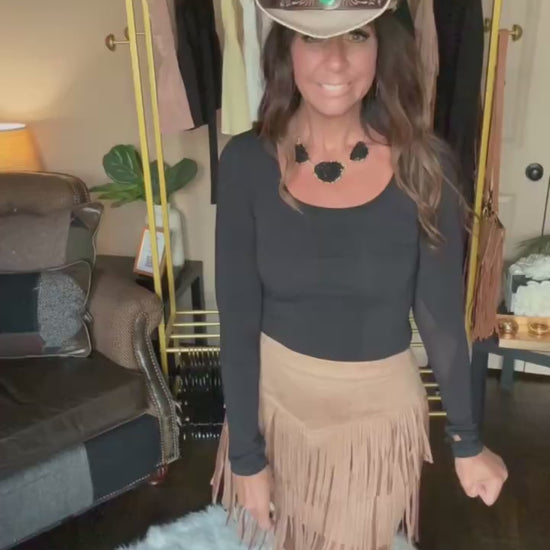 Woman modeling a tan fringe mini skirt in a home boutique.